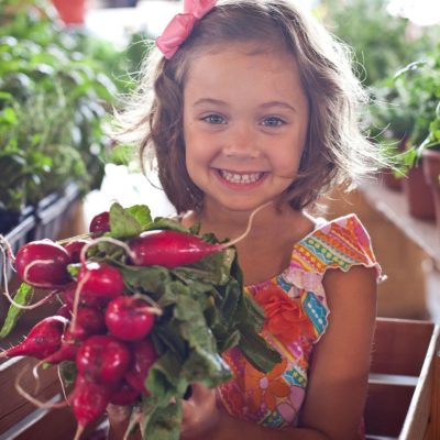 A small girl with a bow in her hair smiling and holding a bunch of radishes at a farmer