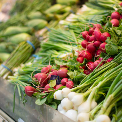 A grocery shelf piled with green onions, red radishes, and cobs of corn