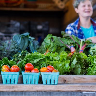 A vendor at a farmers market standing behind a table displaying tomatoes, lettuce and kale