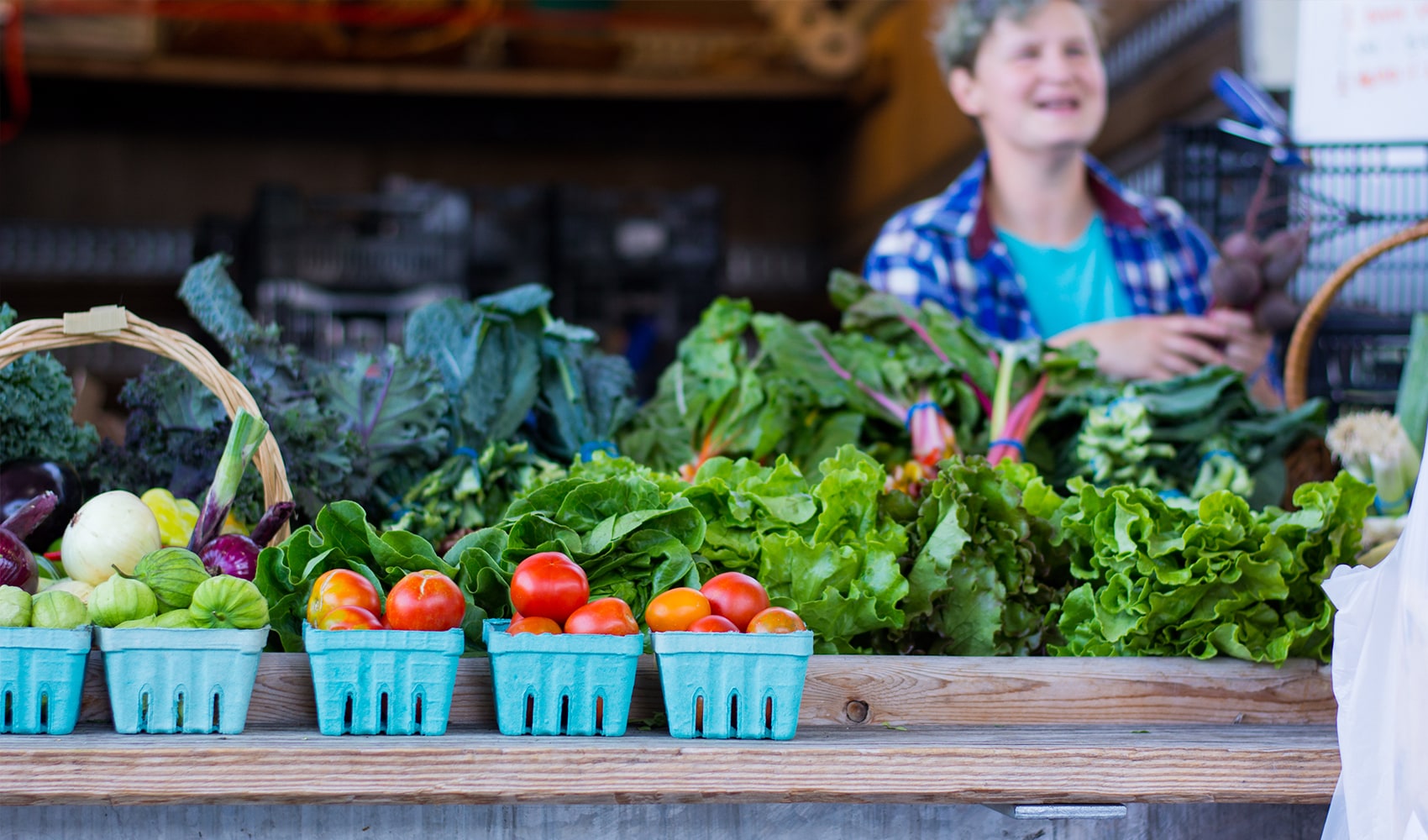 A vendor at a farmers market standing behind a table displaying tomatoes, lettuce and kale