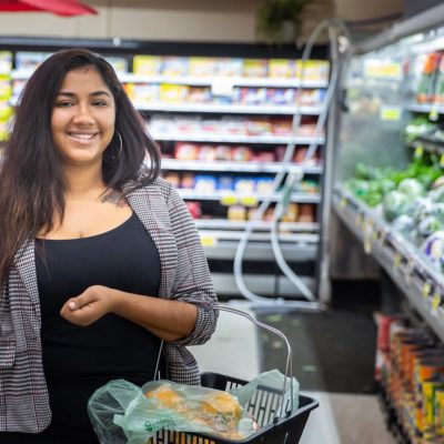 Maxine, a woman with a black shirt and jacket, holding a shopping basket in the produce section of a grocery store
