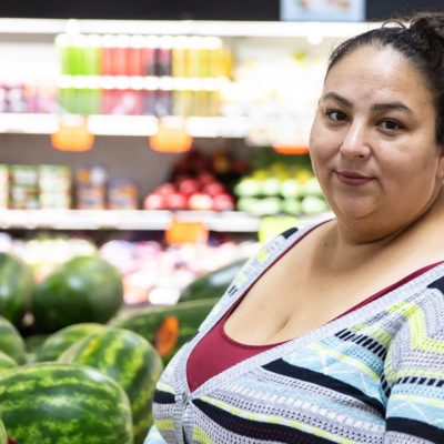 Miroslava, a woman wearing a red shirt and blue sweater standing in a grocery store near watermelons 