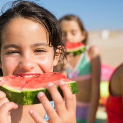 A young girl in a blue swimsuit eating watermelon at the beach with other young girls eating watermelon behind and beside her.
