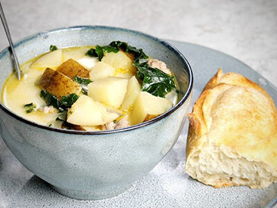 Grey bowl filled with potato and sausage soup on a grey plate with a piece of fresh baguette. 