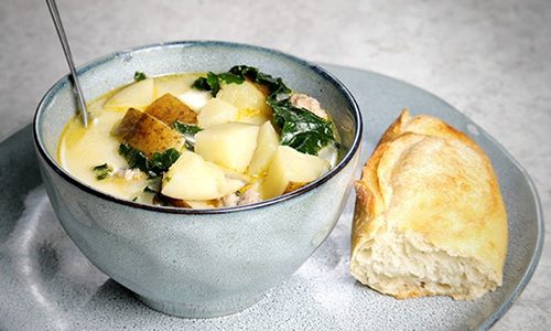 Grey bowl filled with potato and sausage soup on a grey plate with a piece of fresh baguette. 