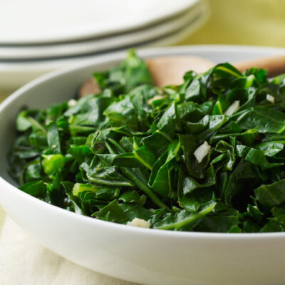 White bowl filled with sauteed collard greens and garlic with a wooden spoon on a table with other plates in the background.