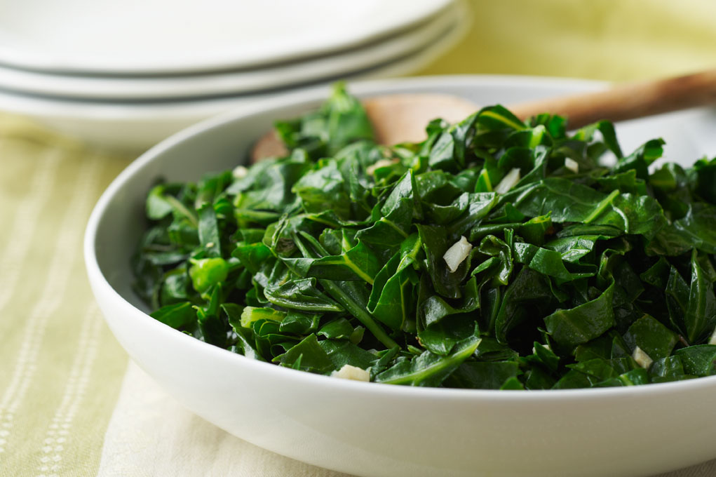 White bowl filled with sauteed collard greens and garlic with a wooden spoon on a table with other plates in the background.