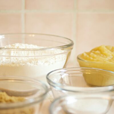 Close up of glass bowls containing baking ingredients including flour, sugar, and applesauce.