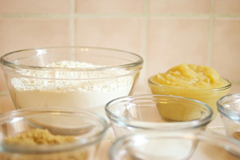 Close up of glass bowls containing baking ingredients including flour, sugar, and applesauce.